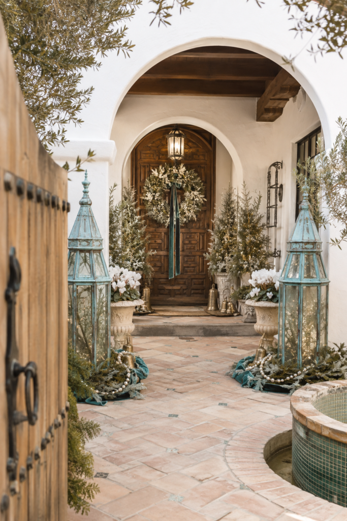 Weathered turquoise lanterns with warm lights flank a Spanish Revival Christmas entry with a frosted wreath brass bells white flowers and pearl garlands
