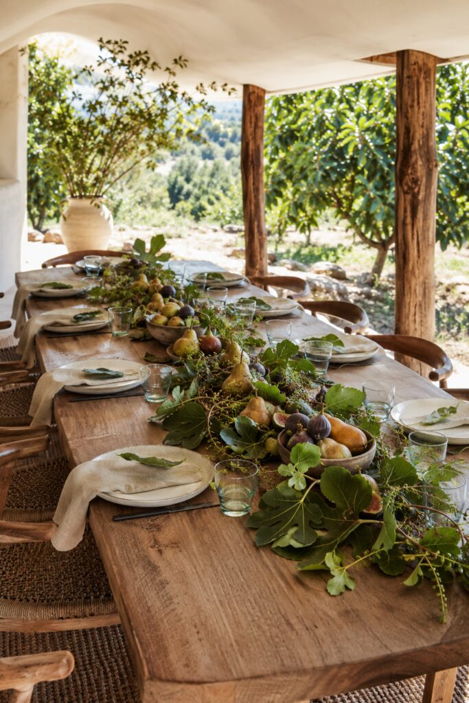 Rustic Thanksgiving table with a flowing fig leaf centerpiece golden pears purple figs ceramic bowls ivory plates oatmeal napkins and pale green glasses