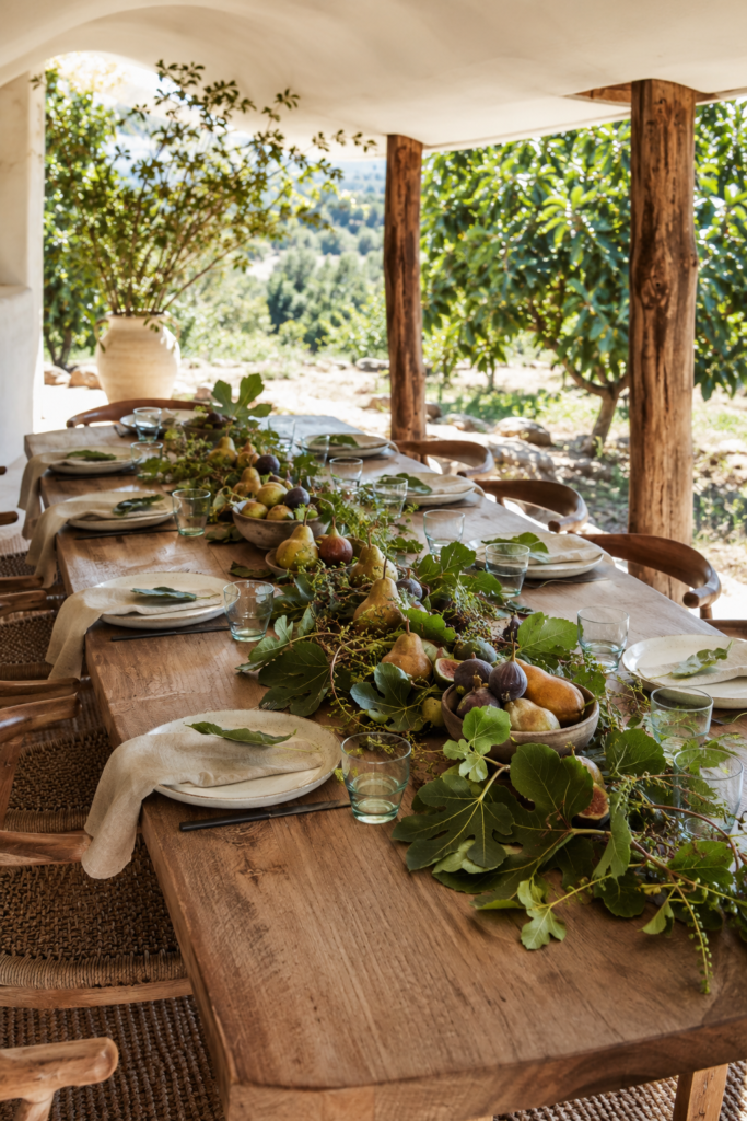 Rustic Thanksgiving table with a flowing fig leaf centerpiece golden pears purple figs ceramic bowls ivory plates oatmeal napkins and pale green glasses
