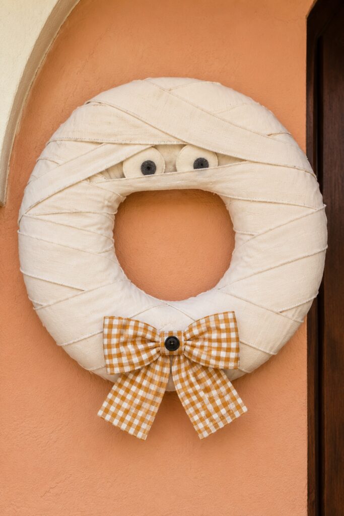 Cream fabric mummy wreath with peeking felt eyes and an ochre gingham bow with a black button on a peach stucco wall