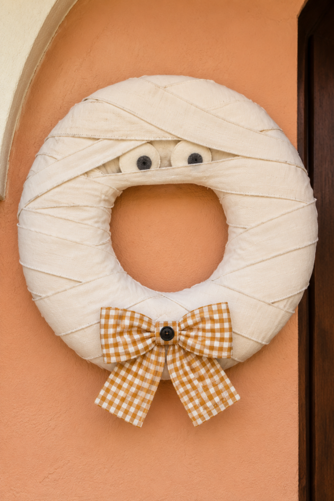 Cream fabric mummy wreath with peeking felt eyes and an ochre gingham bow with a black button on a peach stucco wall