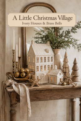 Neutral holiday village with evergreen branches and a linen bow beneath a banner reading A Little Christmas Village Ivory Houses and Brass Bells