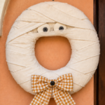 Peekaboo Mummy Wreath text above a Halloween ring wrapped in overlapping ivory strips with soft eyes and a golden checked bow