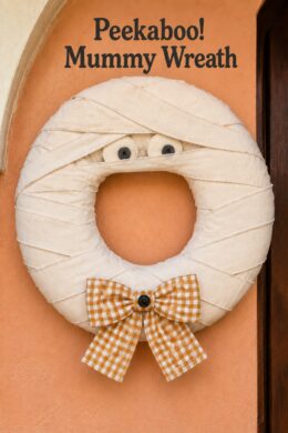 Peekaboo Mummy Wreath text above a Halloween ring wrapped in overlapping ivory strips with soft eyes and a golden checked bow