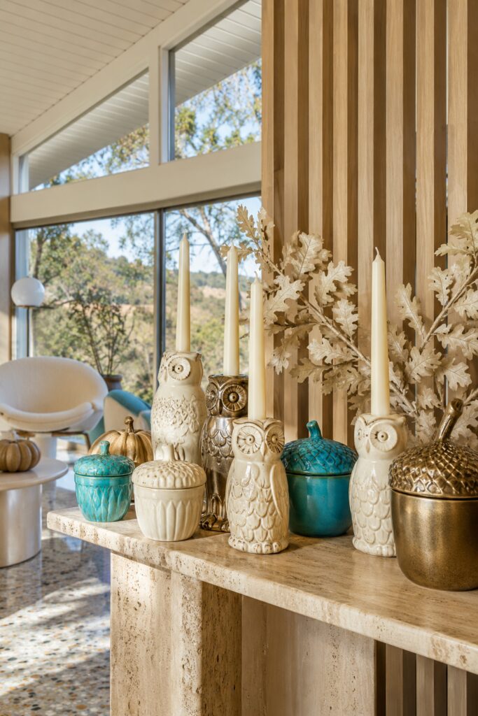 Ivory and gold owl candleholders with turquoise acorn jars and pale oak leaves on a stone console in a sunlit room with a wood slat wall