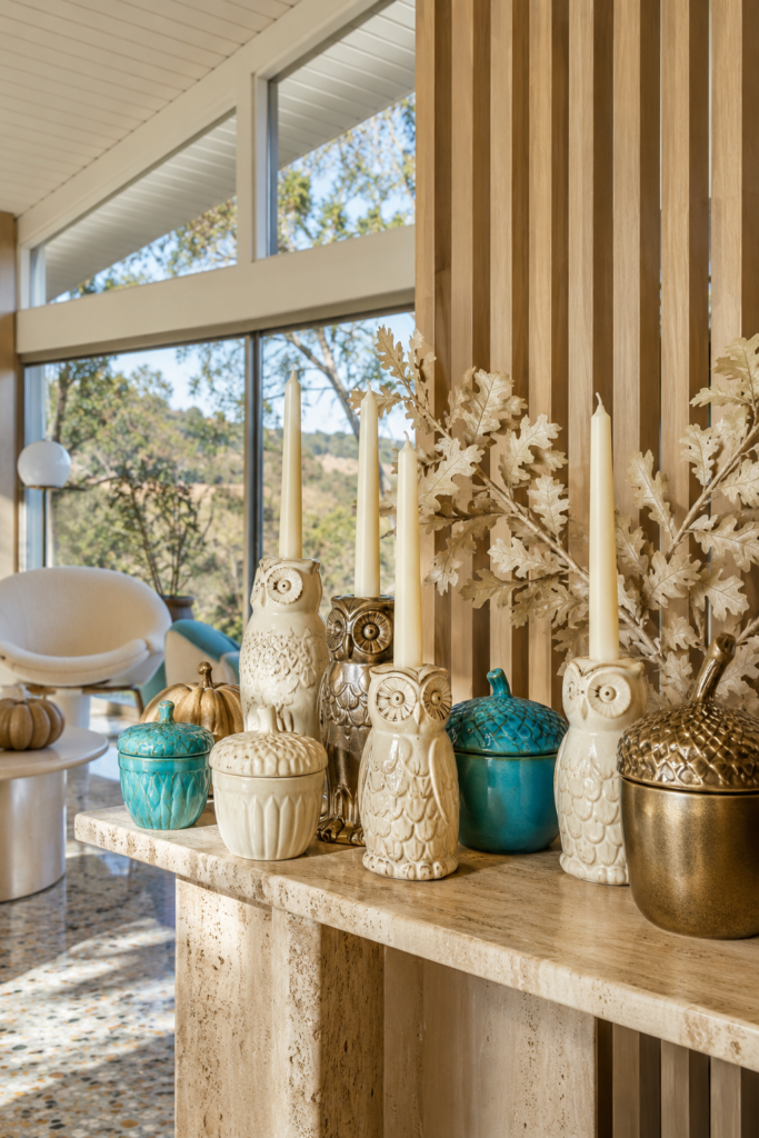 Ivory and gold owl candleholders with turquoise acorn jars and pale oak leaves on a stone console in a sunlit room with a wood slat wall