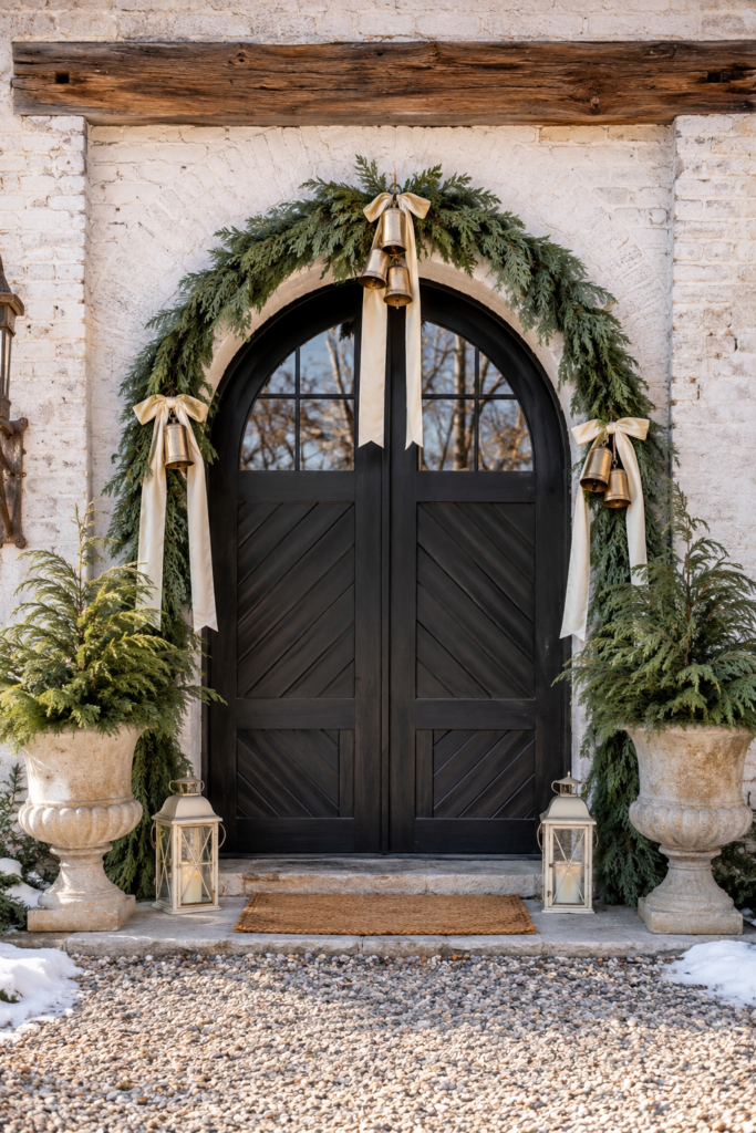 Black arched carriage house doors framed with evergreen garland gold bells and ivory ribbons beside stone urn planters and pale candle lanterns