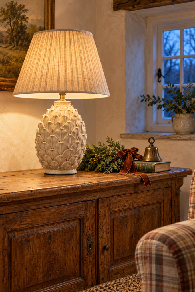Ivory ceramic pinecone lamp with a pleated shade beside cedar greenery cinnamon velvet ribbon and a brass bell on an oak cabinet