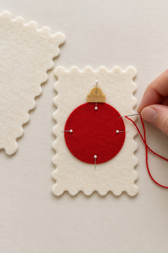 Hand stitching a red felt bauble applique onto one scalloped ivory stamp front beside a separate backing