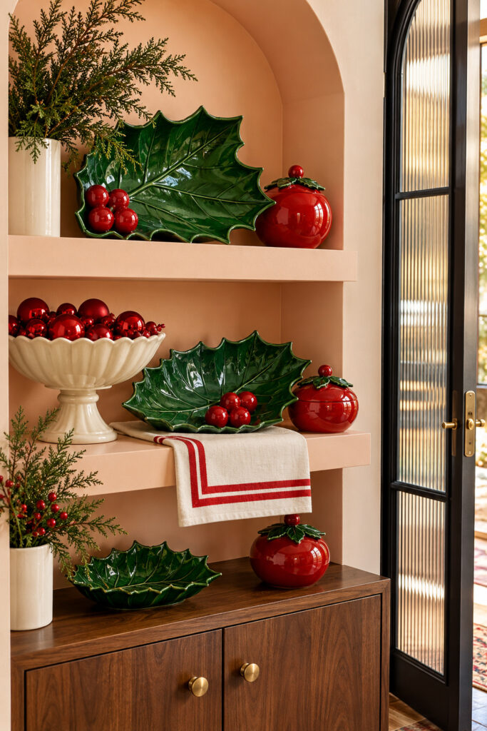 Green holly leaf ceramic dishes and red berry shaped jars arranged on ledges in a peach wall niche