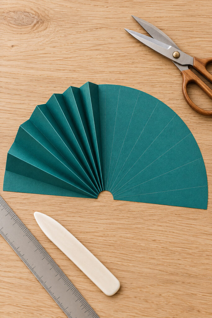 Teal paper semicircle partly folded along radial lines with a small center notch beside a ruler and bone folder