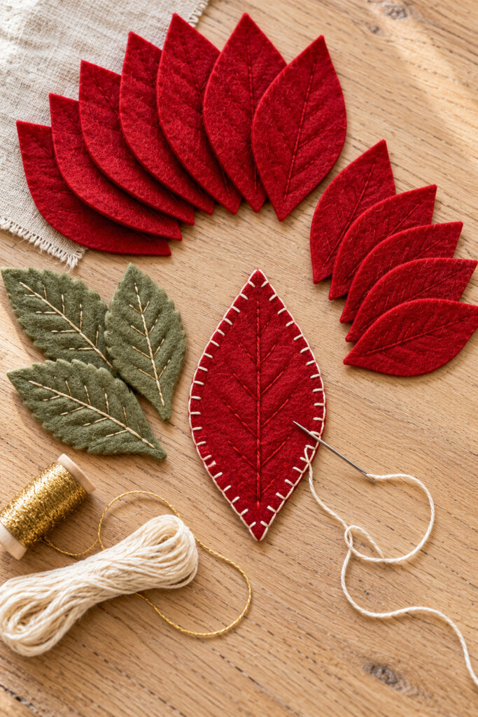 Red felt petal with ivory blanket stitching beside cut red petals sage leaves thread and a needle