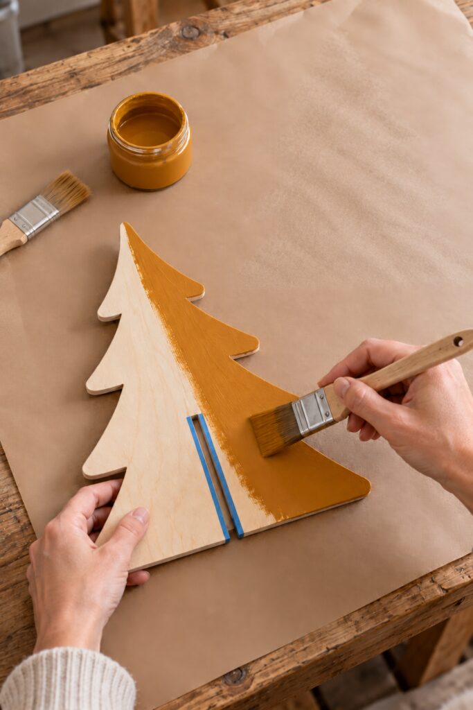 A brush coats a flat plywood tree profile in amber paint beside a taped center slot