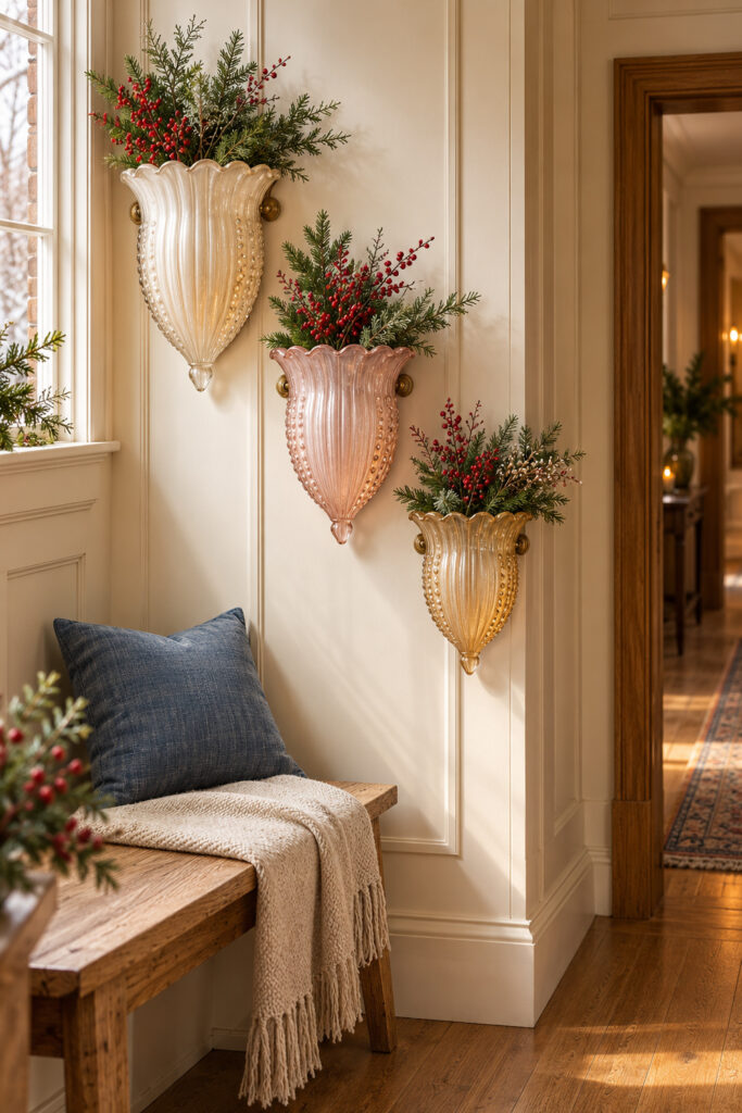 Three scalloped glass wall pocket vases in ivory blush and honey hold evergreen and red berries above an oak hallway bench